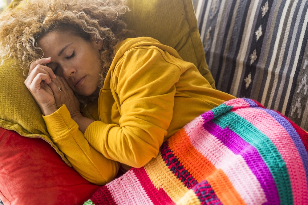 Frau schläft tagsüber erschöpft auf dem Sofa unter einer bunten Decke