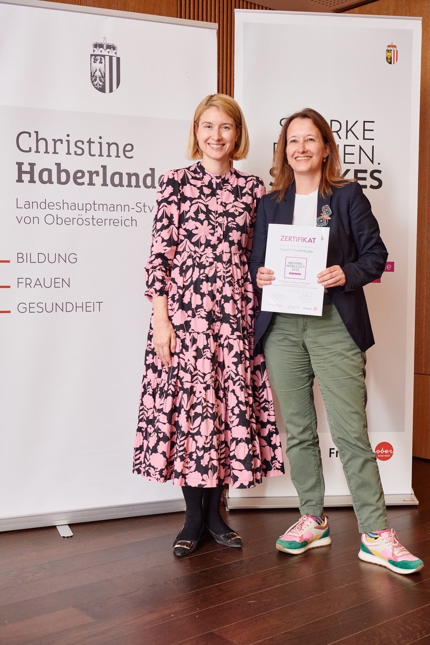 Dr. Gerhilt Kusatz und Landeshauptmann-Stellvertreterin Mag. Christine Haberlander stehen lächelnd nebeneinander vor Roll-up-Bannern, Dr. Kusatz hält ein Zertifikat der #BeWirkwerkstatt 2030 in der Hand